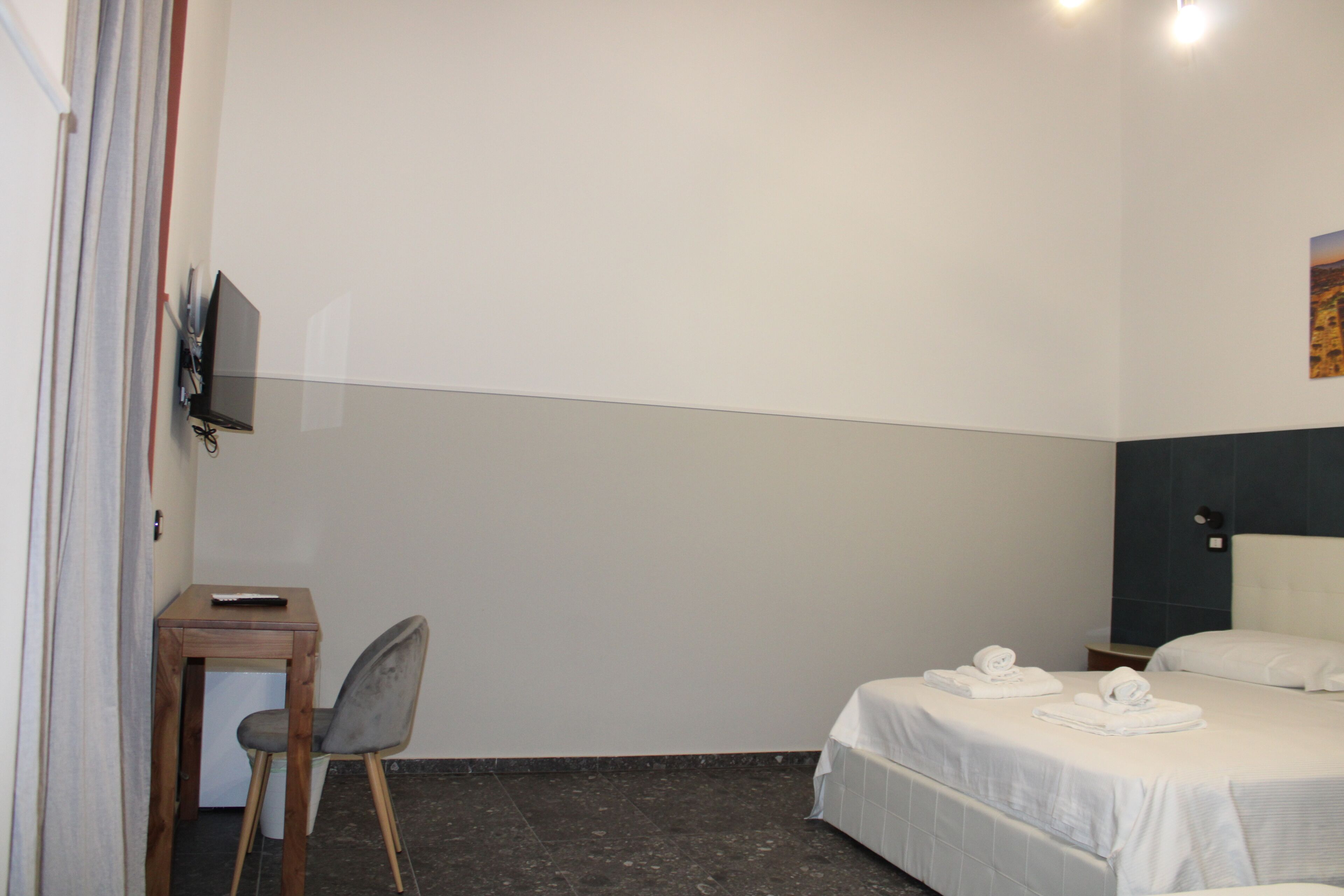 A hotel room with a bed, a desk with a chair, a TV mounted on the wall, and a small trash bin.