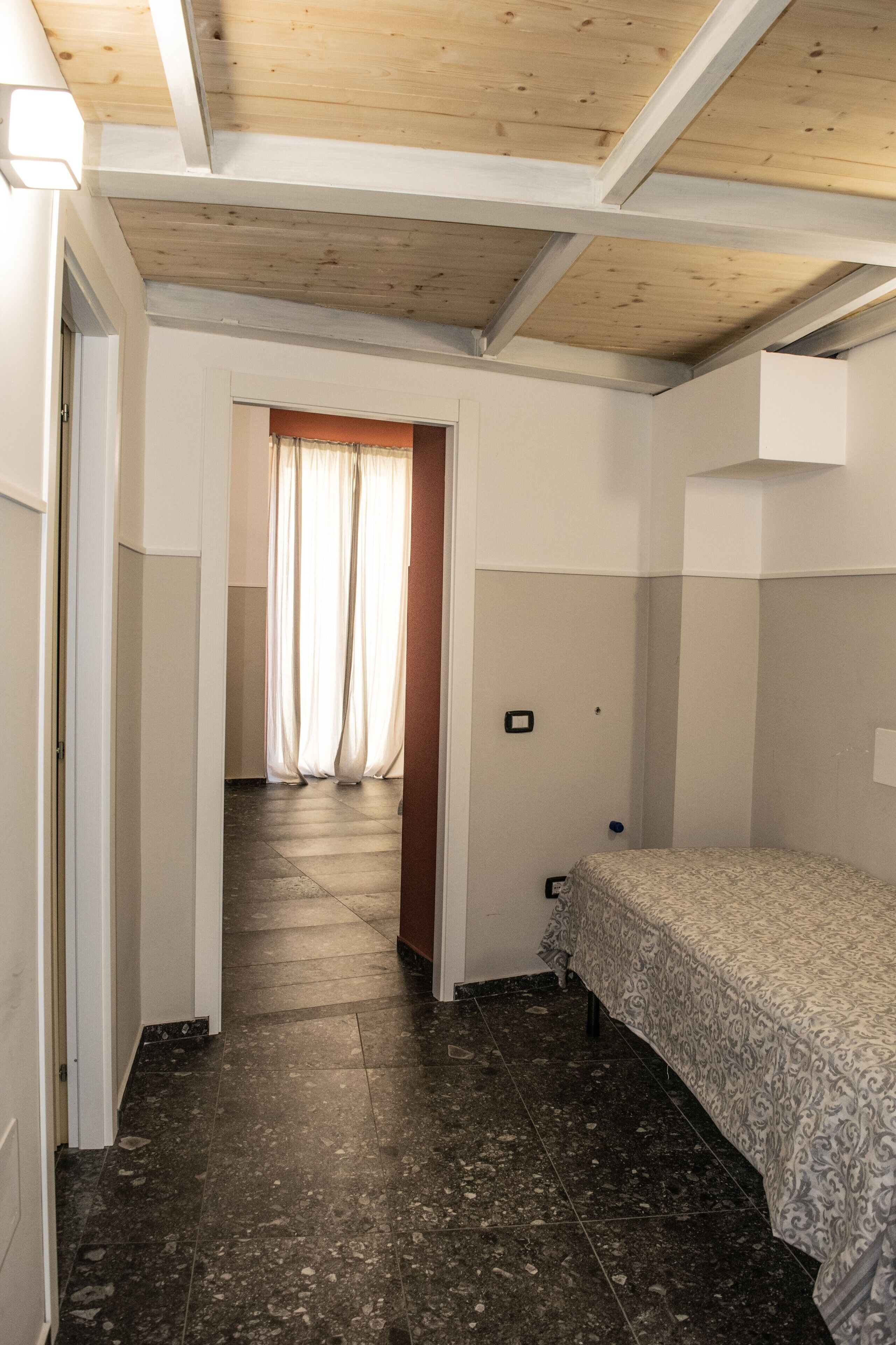A narrow hallway leading to a bedroom with a bed and a window with curtains.