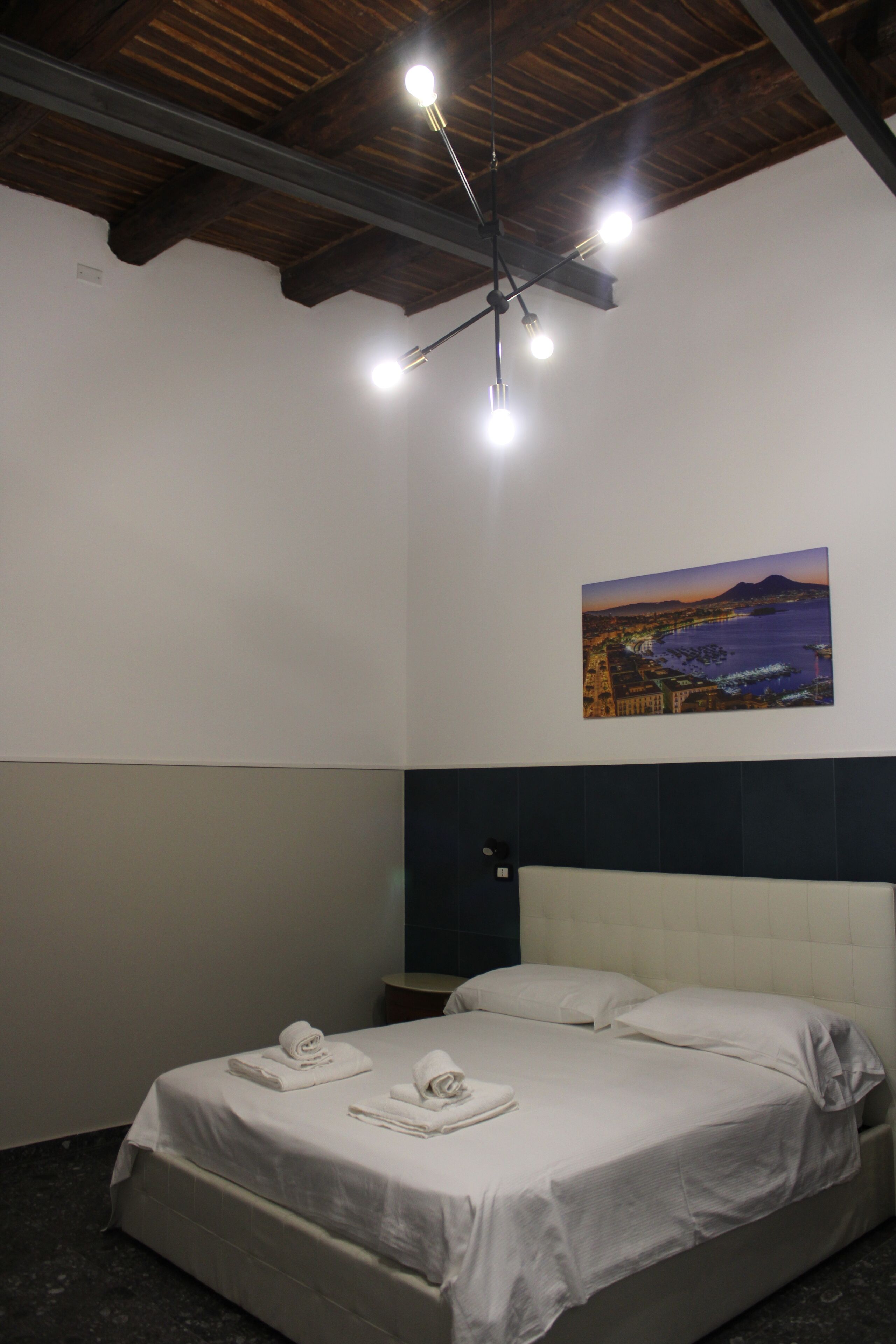 Una habitación de hotel con una cama, un cuadro de un paisaje y un techo con vigas y luces a la vista.