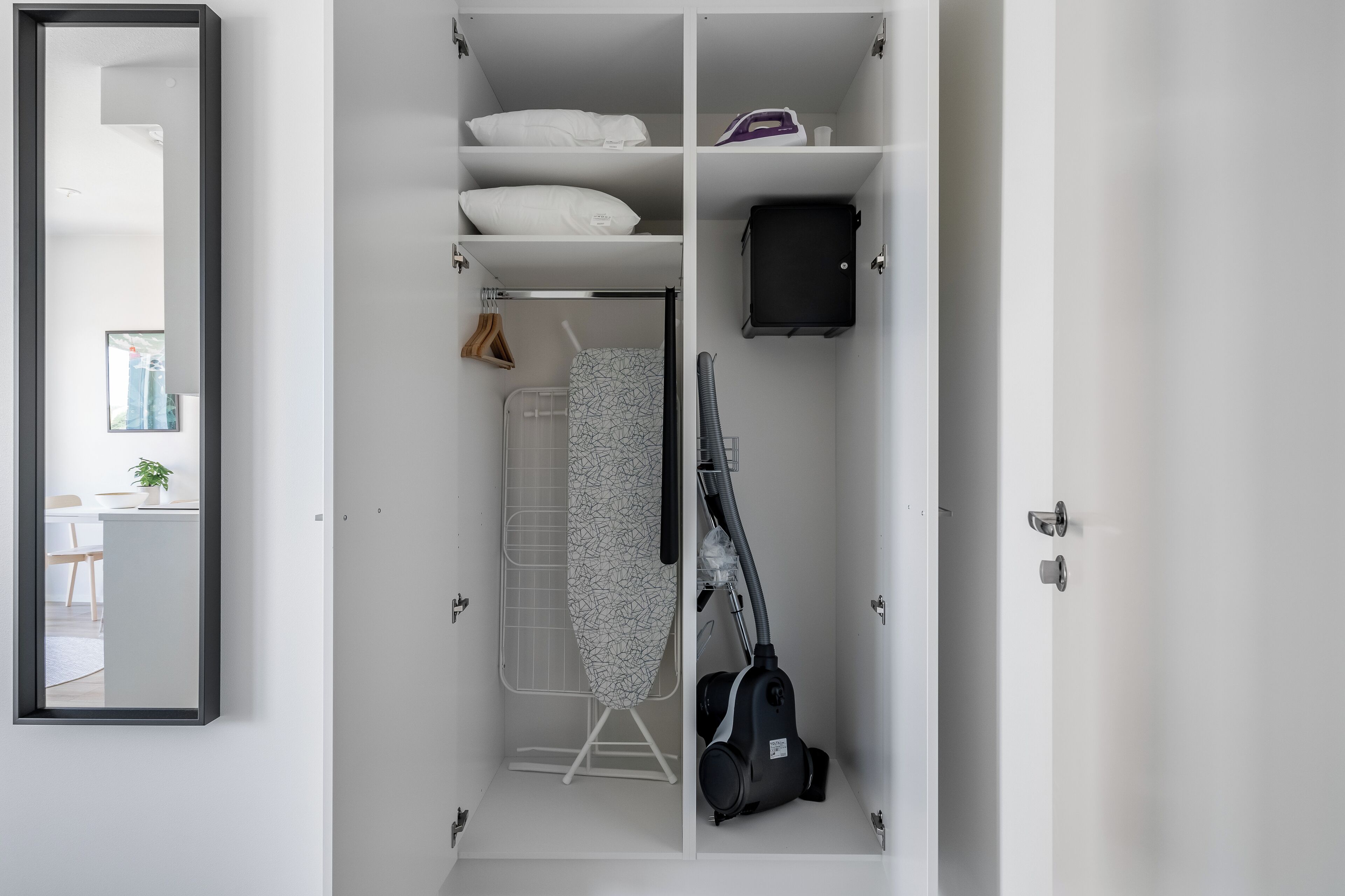 A white wardrobe with a mirror, shelves with pillows, a hanging rack with clothes, and a vacuum cleaner.