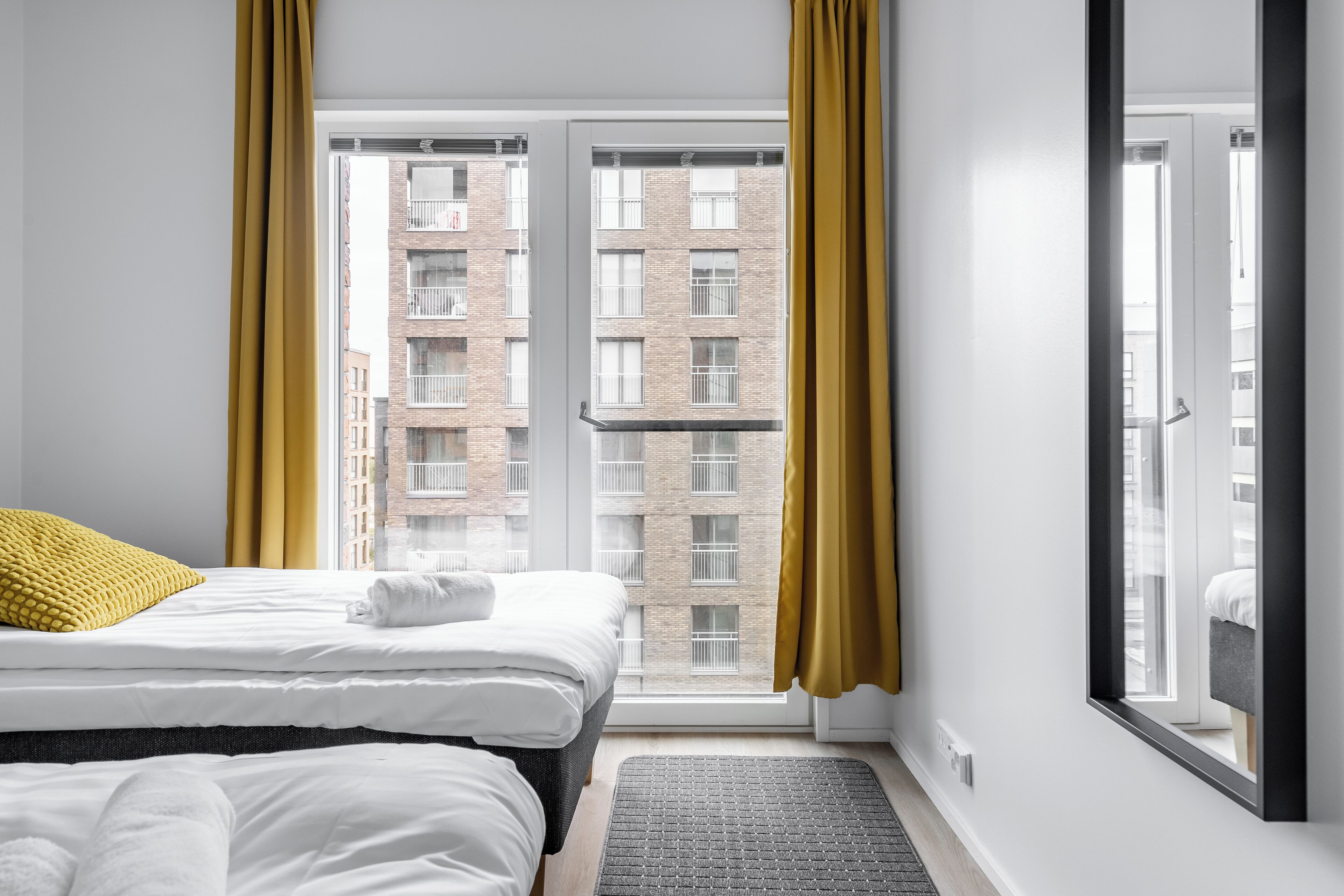 Una habitación de hotel con una cama, un ventanal con cortinas amarillas y vista a un edificio de ladrillo.