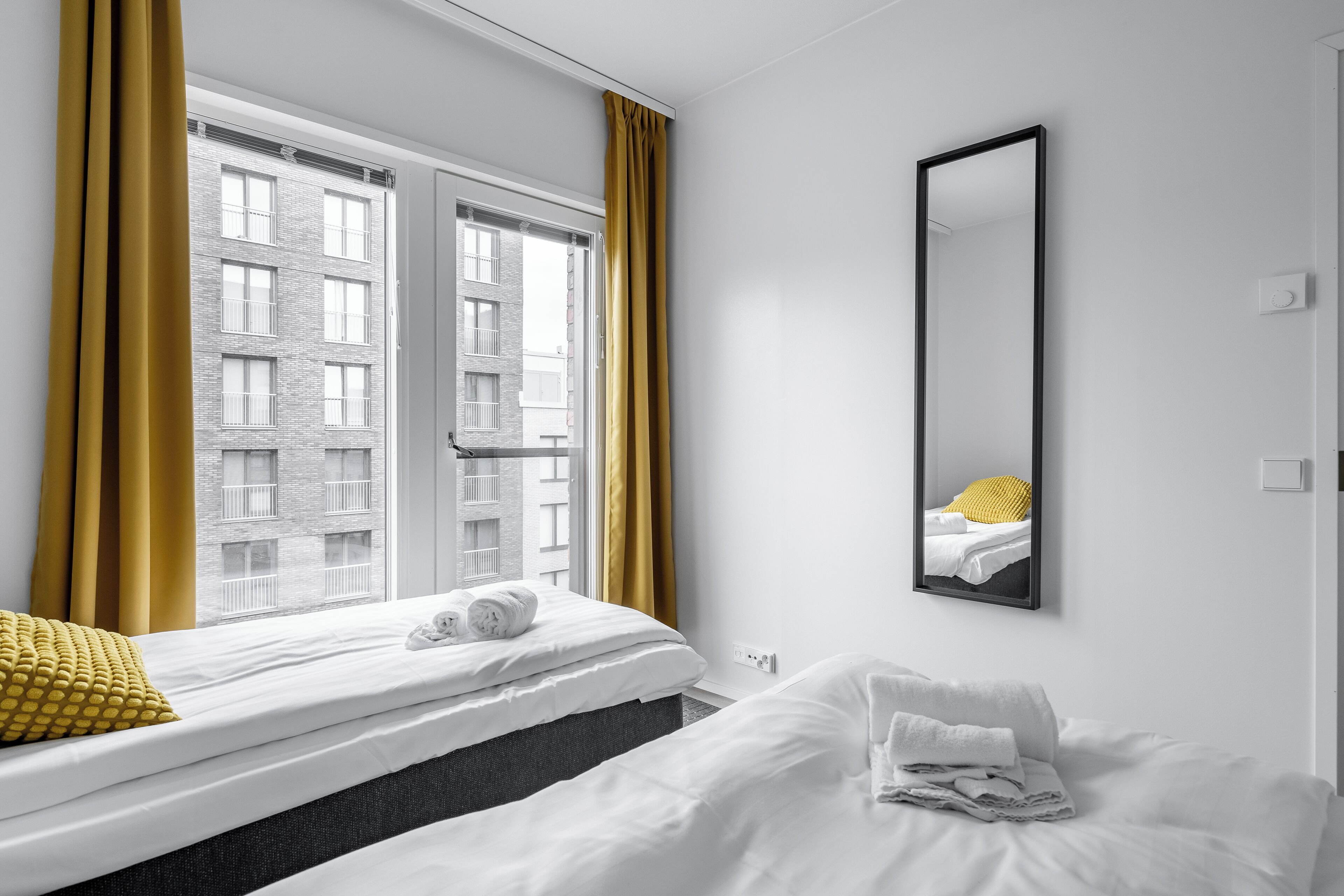 A hotel room with two beds, a large window with curtains, a mirror, and a view of a building outside.
