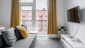 A modern living room with a grey sofa, mustard curtains, a flat-screen TV, and a view of a red brick building through a large window. - Hiisi Homes Turku Herttuankulma (Turku)