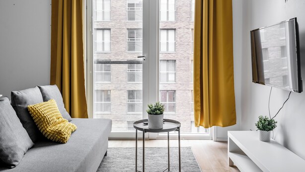 A modern living room with a grey sofa, yellow cushions, a small round table with a plant, and a TV mounted on the wall. - Hiisi Homes Turku Herttuankulma (Turku)