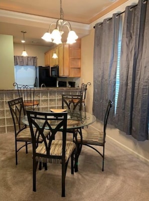 A dining area with a round glass table and dark wooden chairs, a kitchen in the background, and a curtain on the right. - Lolly Gris-Gris (Hot Springs)