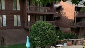 A swimming pool with lounge chairs and umbrellas, surrounded by a wooden deck and a brick building in the background. - Lolly Gris-Gris (Hot Springs)