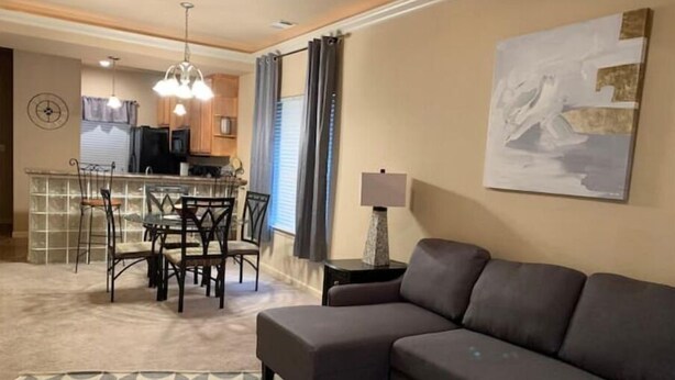 A living room with a grey sofa, a patterned rug, a dining area with a table and chairs, a kitchen in the background, and a painting on the wall. - Lolly Gris-Gris (Hot Springs)