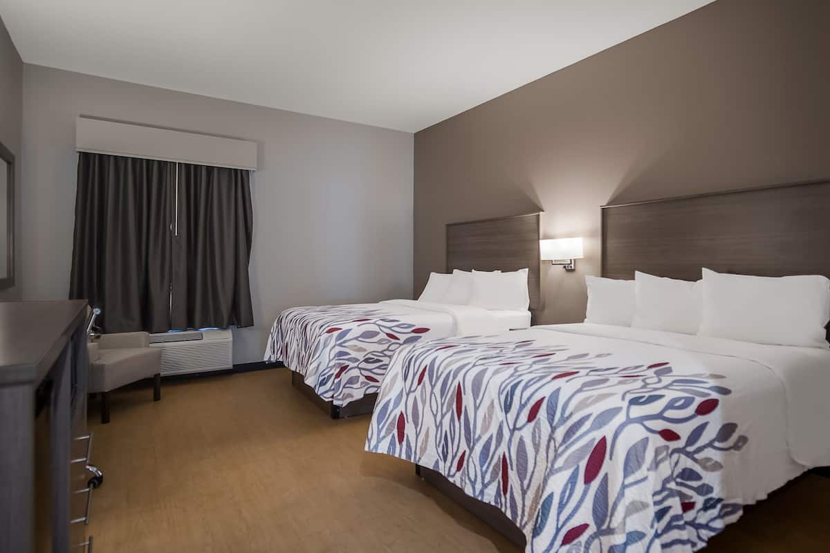 Ett hotellrum med två sängar, ett skrivbord, en stol, ett fönster med gardiner och en vägglampa.