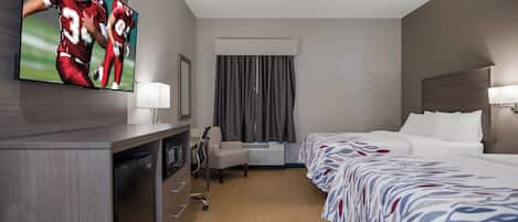 Een hotelkamer met een bed, een bureau met een computer, een aan de muur bevestigde televisie en een raam met gordijnen.