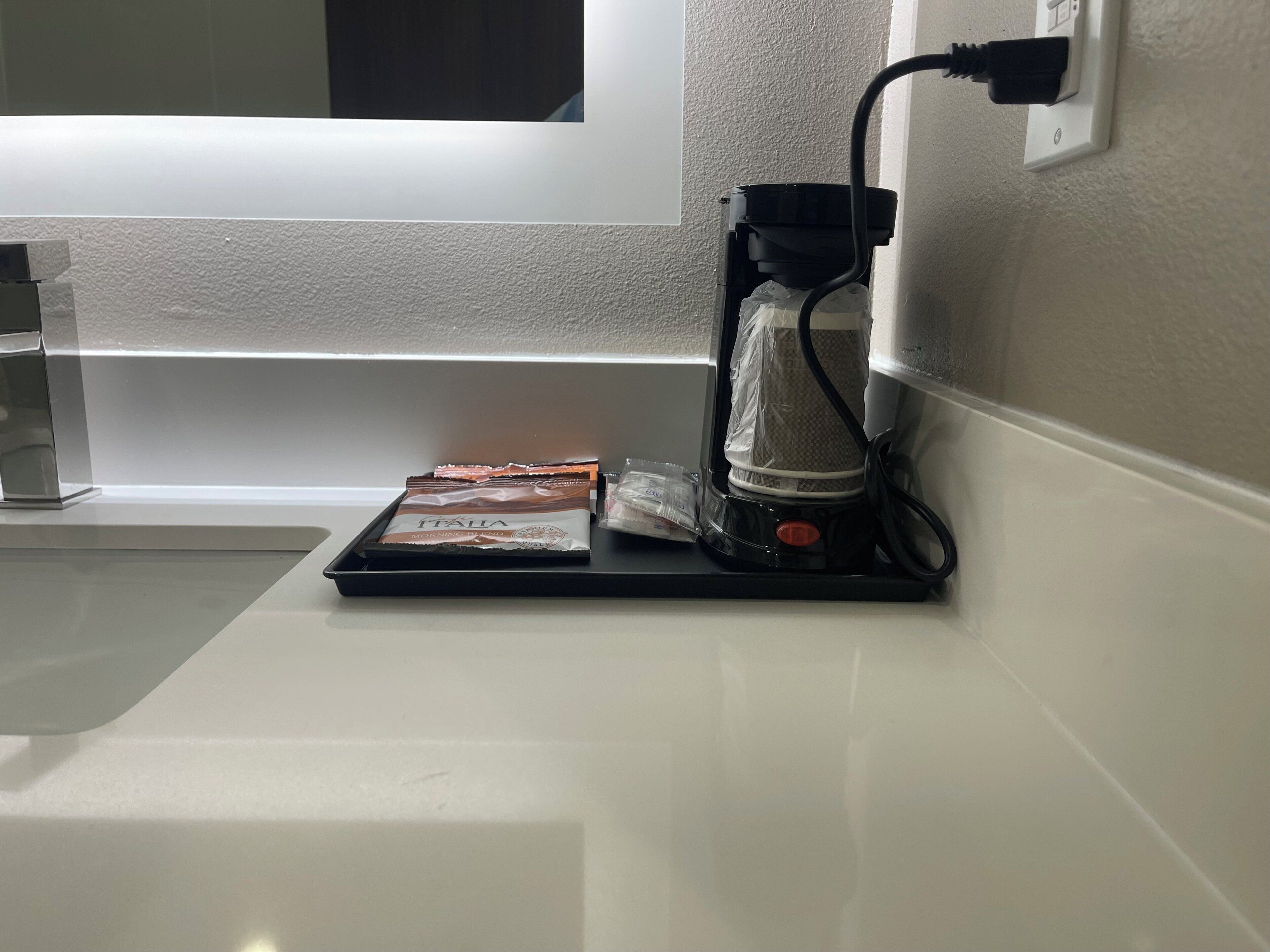 A coffee machine with a tray of coffee pods and a bag of sugar on a white countertop near a wall socket.