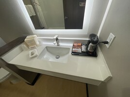 Un baño con lavamanos blanco, un espejo encima y una cafetera sobre la encimera.