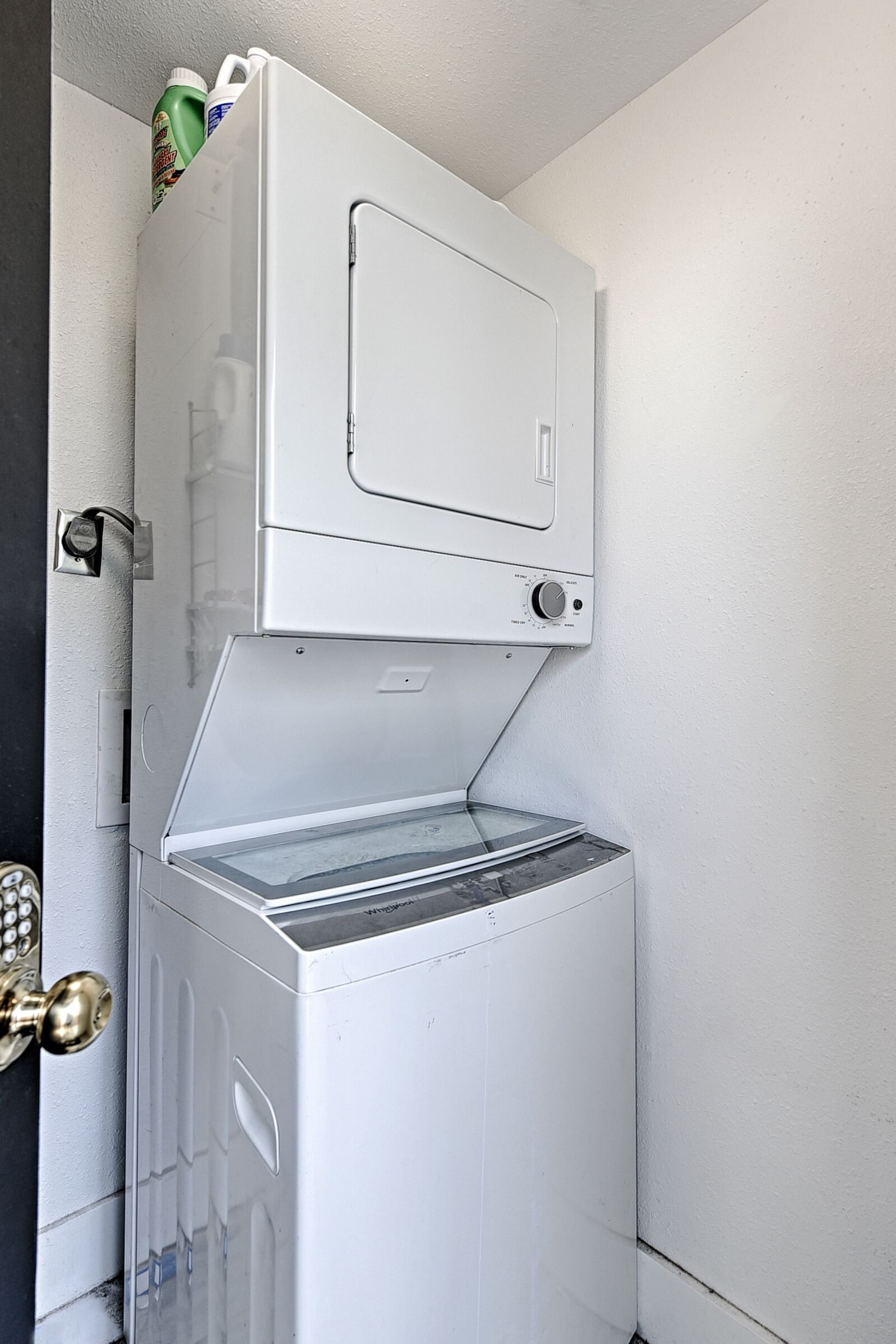 A compact laundry room with a stacked washer and dryer, a door with a brass handle, and a small shelf above the dryer holding cleaning supplies.