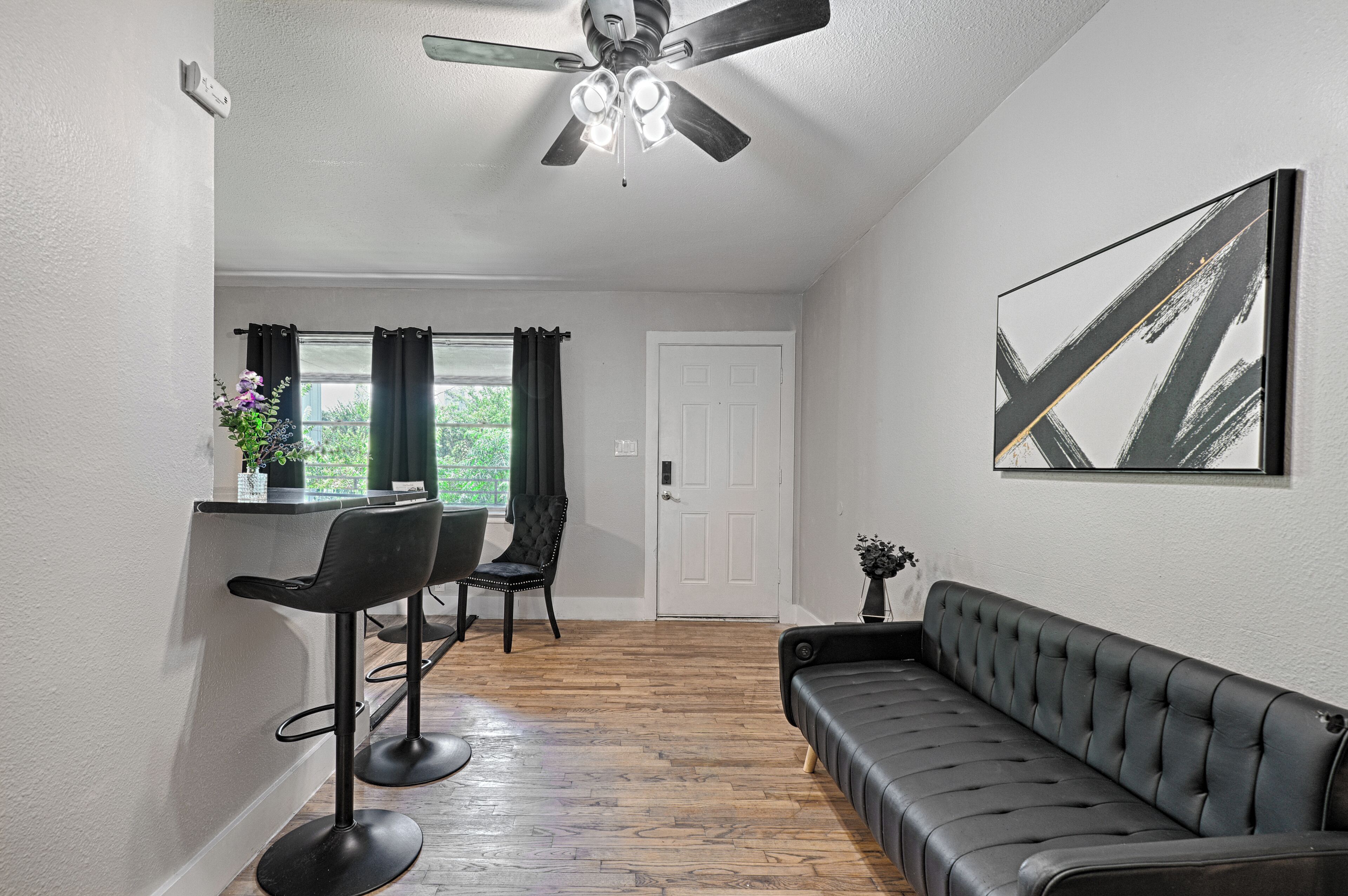 A modern living room with a black leather sofa, a dining area with a bar stool, and a ceiling fan.