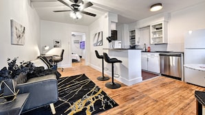 Un salon moderne comprenant un canapé, deux tabourets de bar, un ventilateur de plafond et un coin cuisine avec des meubles blancs et des appareils électroménagers en acier inoxydable.