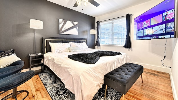 Une chambre à coucher moderne avec un lit, un ventilateur de plafond, un téléviseur fixé au mur et une fenêtre avec des rideaux.