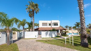 A modern two-story house with a stone pathway leading to a pool area, surrounded by palm trees and a clear blue sky.
