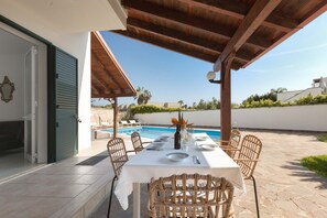 An outdoor dining area with a table set for four, surrounded by wicker chairs, under a wooden pergola, with a pool in the background. - Villa Sunbeach by BarbarHouse (Torre dell'Orso)