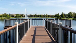A wooden deck with railings leading to a body of water, surrounded by greenery and houses.