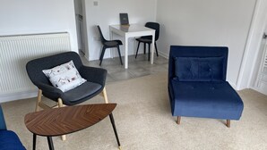A living room with a dark grey armchair, a blue sofa, a wooden coffee table, and a white dining table with two chairs.