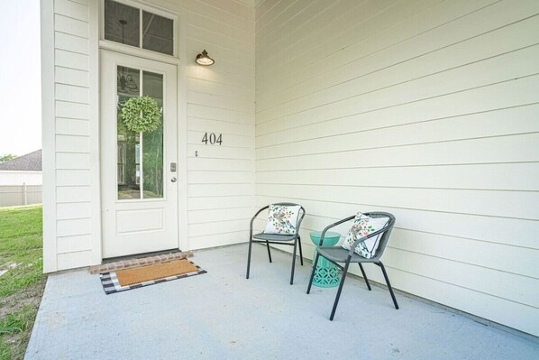 Een wit huis met het nummer 404 op de deur, twee metalen stoelen met bloemenkussens, en een kleine groene prullenbak.
