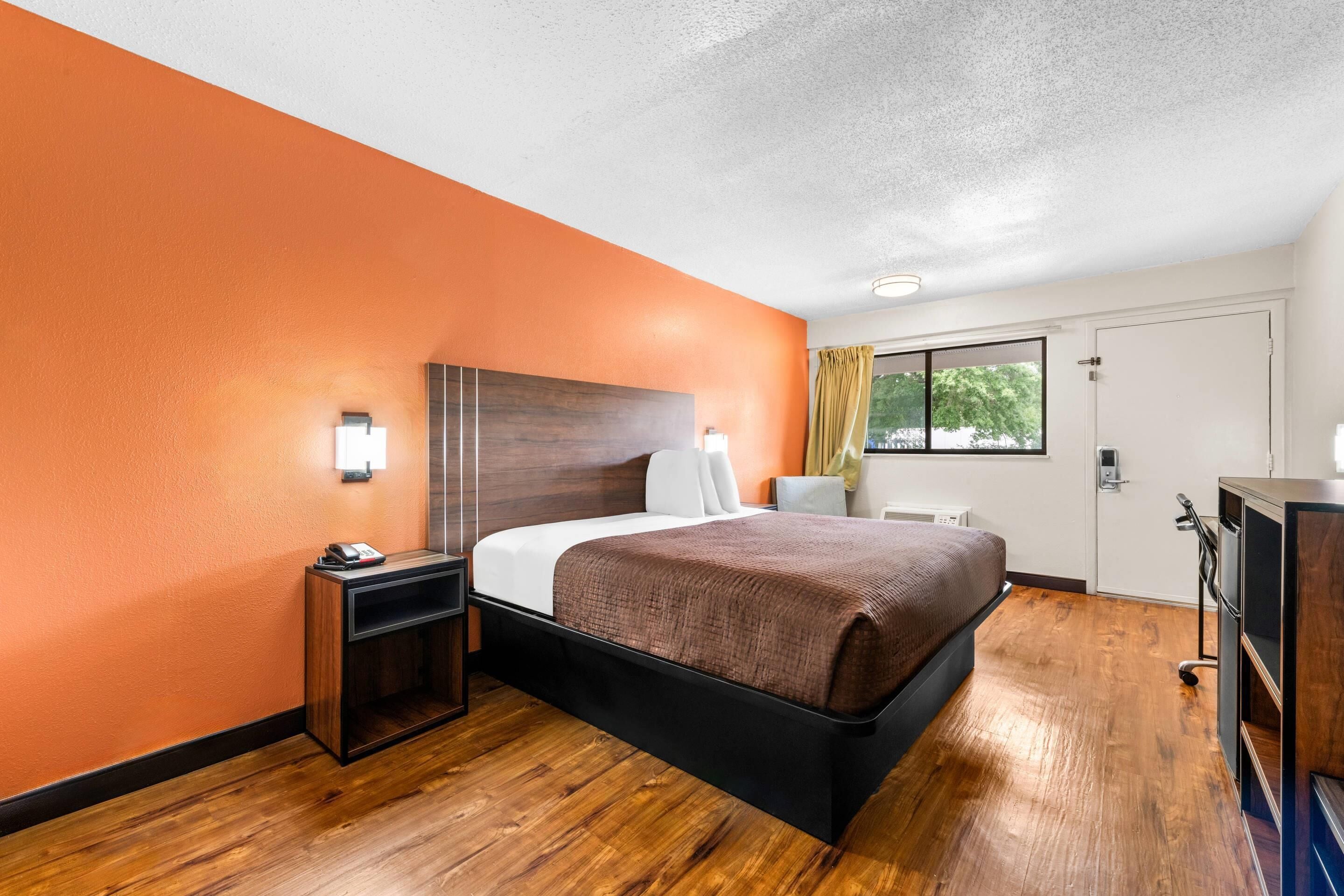 Habitación de hotel con piso de madera, una cama con manta marrón, una mesita de noche, un escritorio y una ventana con cortinas.