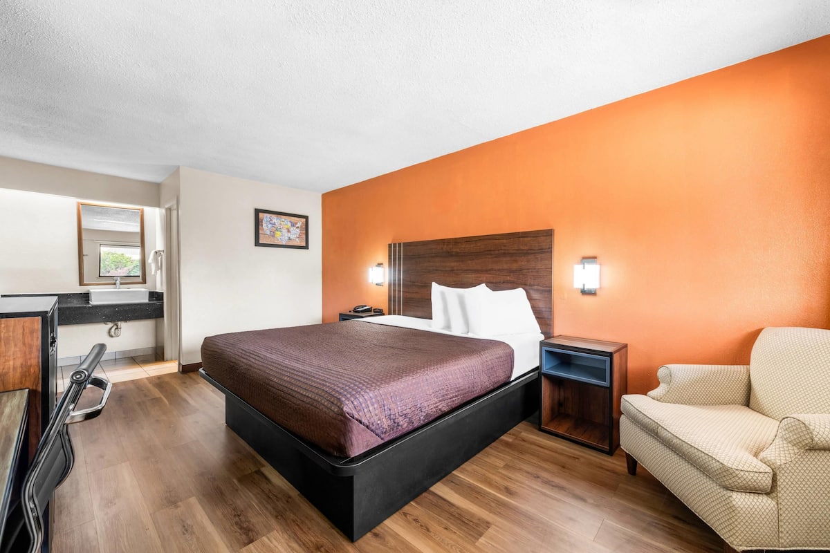 Une chambre d’hôtel avec un lit, une table de chevet, une chaise, un bureau et une salle de bain visible à travers une porte ouverte.