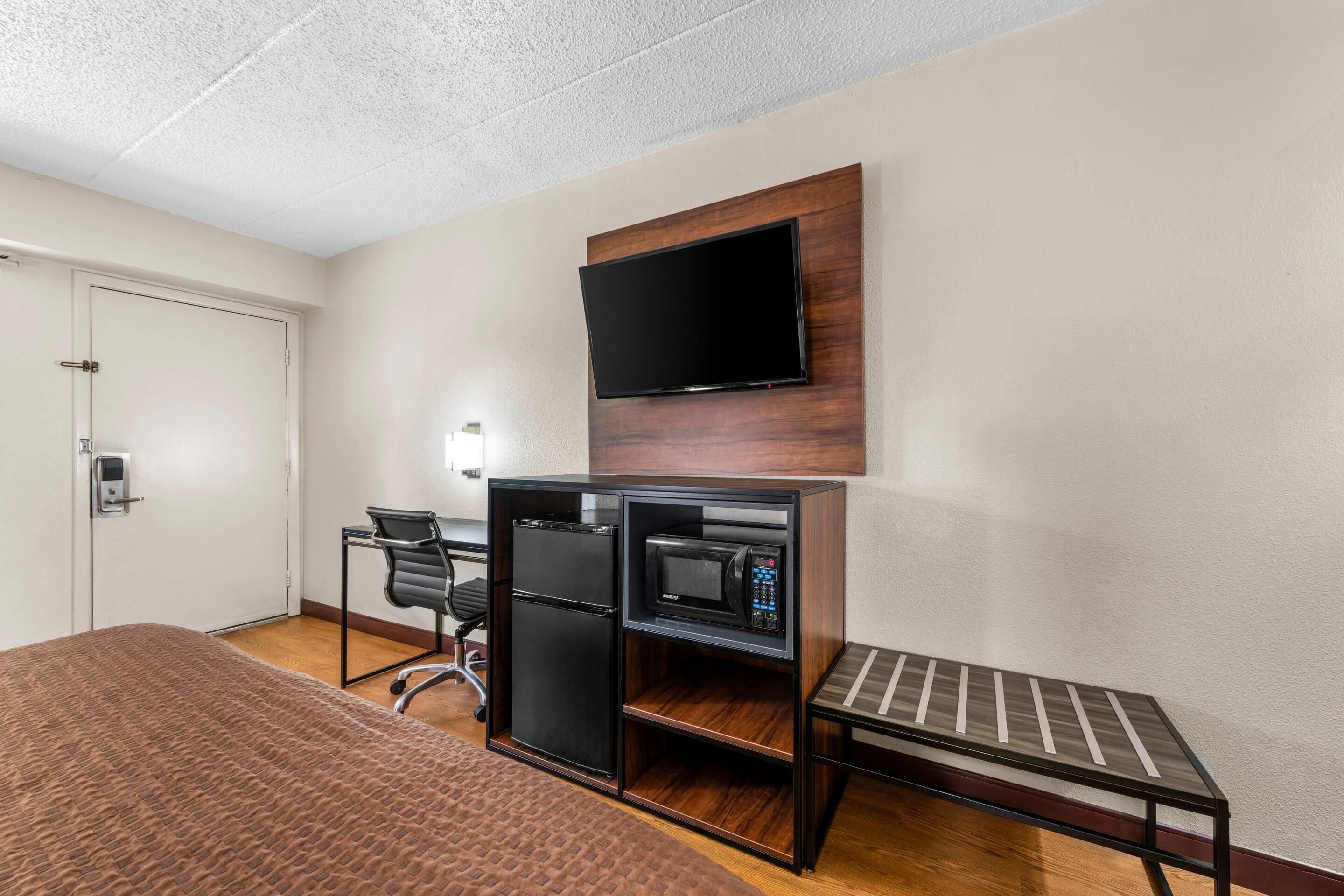Ein kompaktes Hotelzimmer mit Bett, Schreibtisch mit Computer und Mikrowelle, an der Wand befestigtem Fernseher und einer Sitzbank.