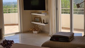 Quarto com televisão de tela plana na parede, cama com manta dobrada, mesa pequena com um vaso e varanda com vista para vegetação e edifícios.