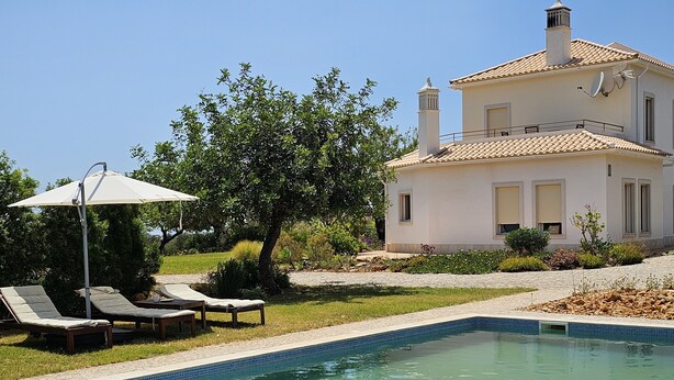 Uma área de piscina com espreguiçadeiras e um guarda-sol, rodeada por um jardim e uma casa com telhado de azulejos.