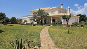 Uma casa de dois andares com telhado de telhas, um caminho de pedra que leva à entrada e um jardim bem cuidado com diversas plantas e uma pequena árvore.