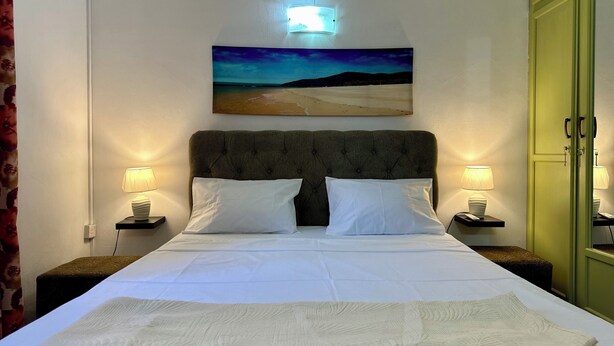 A neatly made bed with white linens and a folded blanket, two bedside lamps, a wall-mounted painting of a beach scene, and a green door to the right.