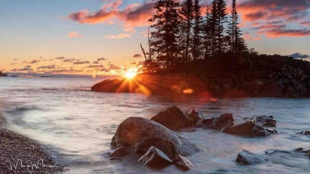 A sunset over a rocky shoreline with trees in the background. - Iconic Knotty Pine Cabin in Charming Grand Marais (Grand Marais)