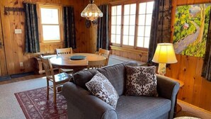 A cozy room with wooden walls, a round dining table, a gray sofa with patterned cushions, and a lamp. - Iconic Knotty Pine Cabin in Charming Grand Marais (Grand Marais)
