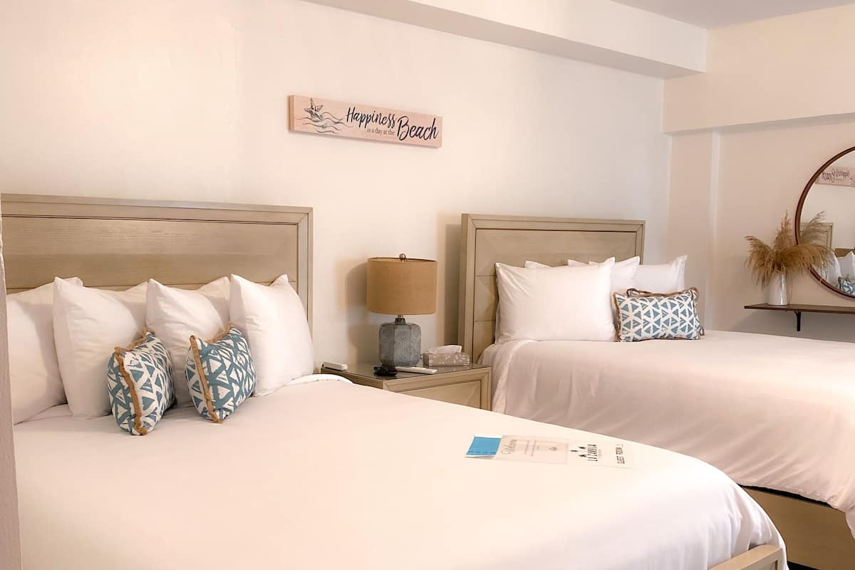 A hotel room with two beds, each with white bedding and patterned pillows. There is a nightstand with a lamp and a decorative sign on the wall.