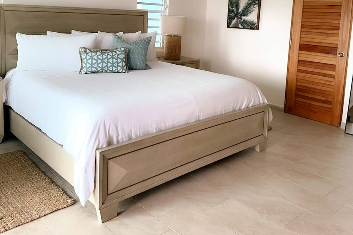 A bedroom with a large bed, a wooden headboard, a bedside lamp, a framed picture on the wall, and a wooden door.