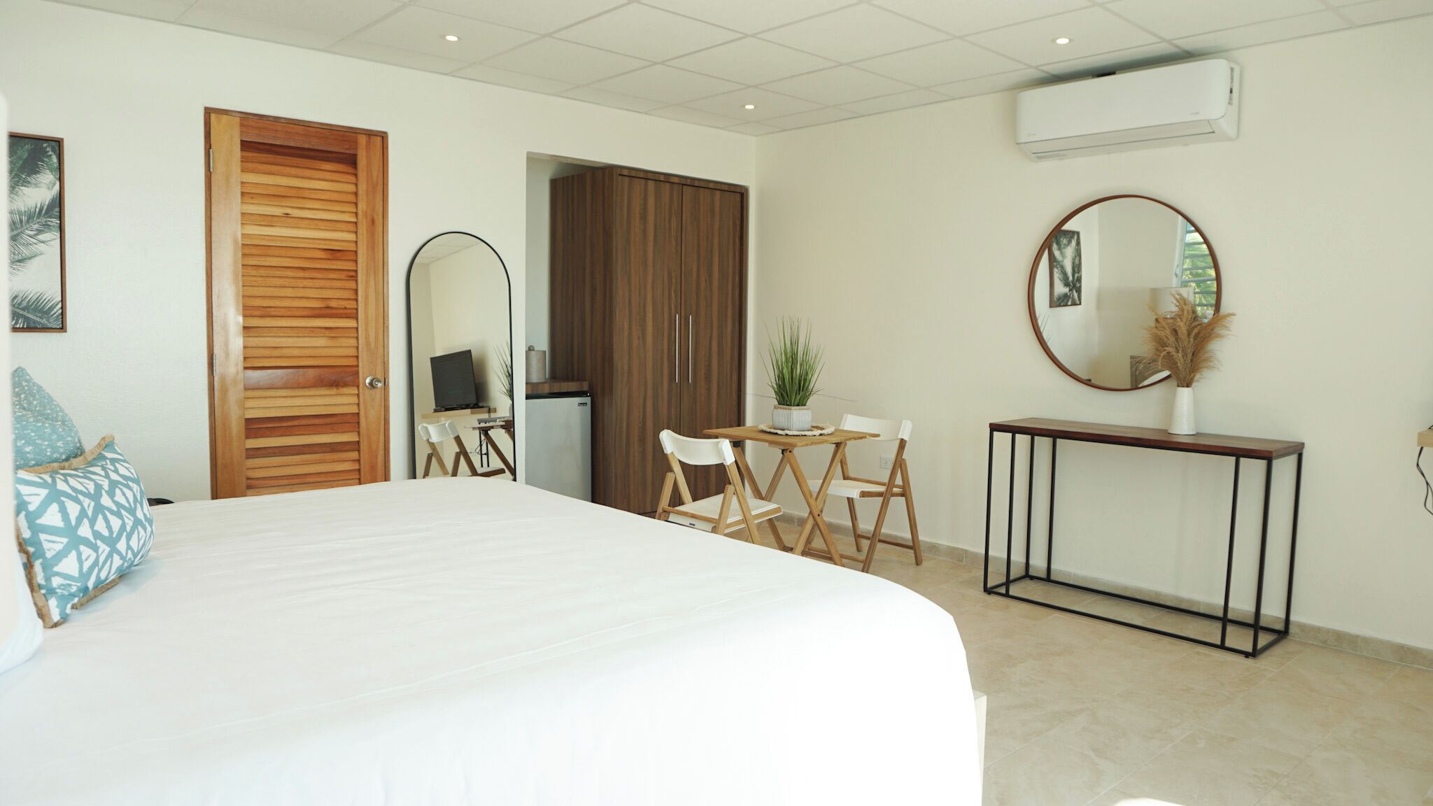 Un dormitorio con cama, escritorio, silla, mesita, espejo y unidad de aire acondicionado.