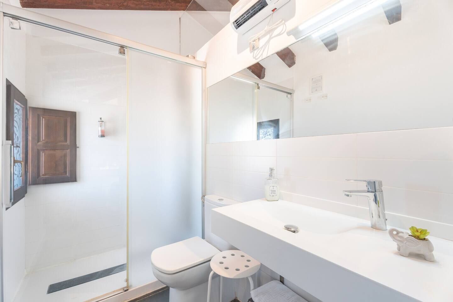 Ein modernes Badezimmer mit Duschkabine aus Glas, einer weißen Toilette, einem Waschbecken mit Spiegel und einem kleinen Hocker.