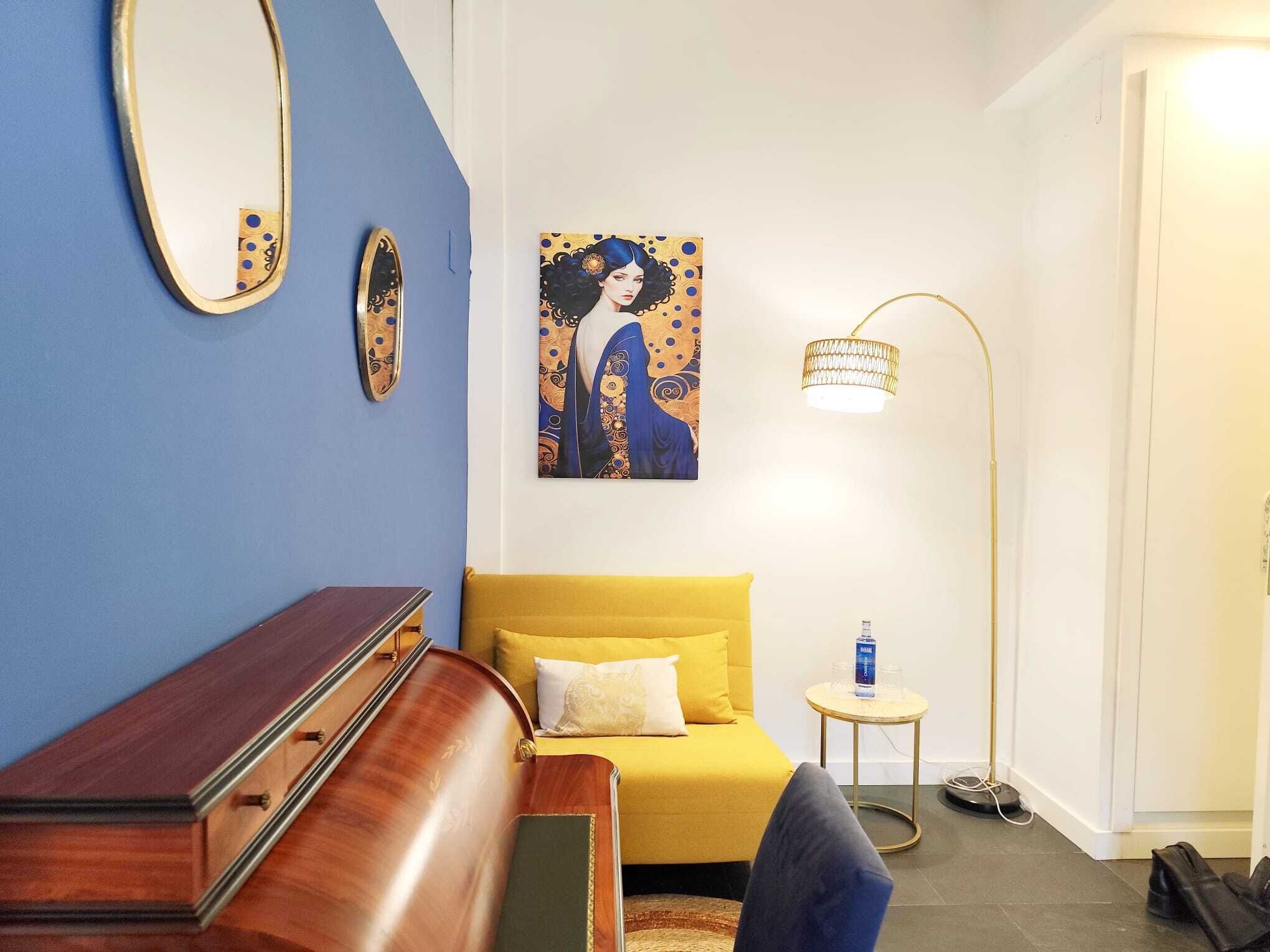 Een kamer met een blauwe wand, een gele bank, een houten bankje, een schilderij, een vloerlamp en een kleine ronde tafel met een fles en een glas.