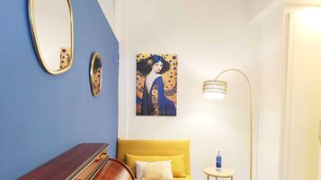 Een kamer met een blauwe wand, een gele bank, een houten bankje, een schilderij, een vloerlamp en een kleine ronde tafel met een fles en een glas.