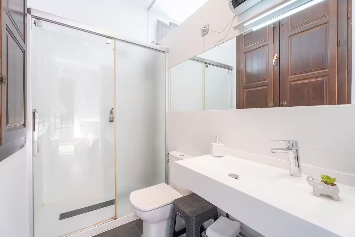 A modern bathroom with a glass shower enclosure, a white toilet, a white sink with a mirror above, and wooden cabinets.