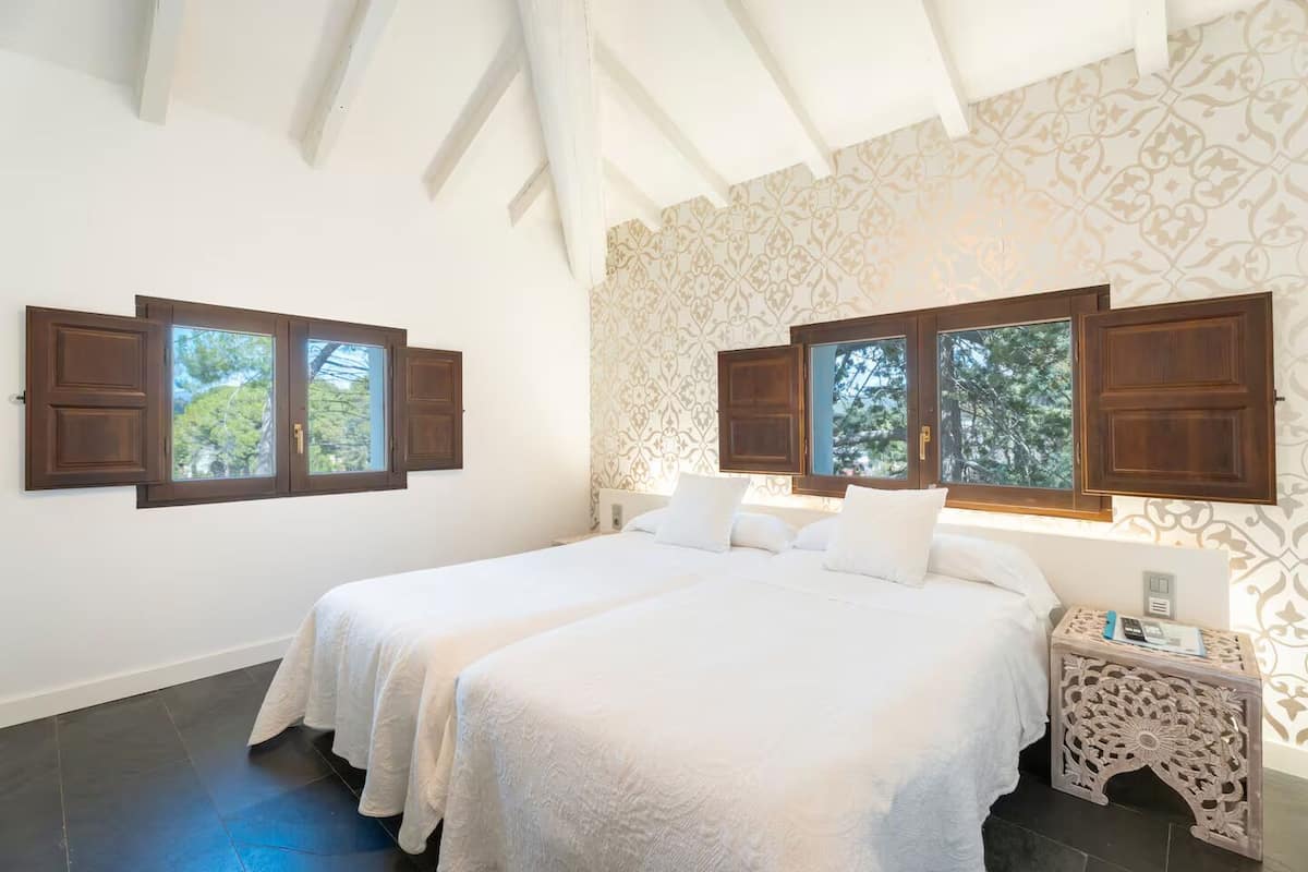 A bedroom with two beds, large windows with wooden shutters, patterned wallpaper, and a decorative nightstand.