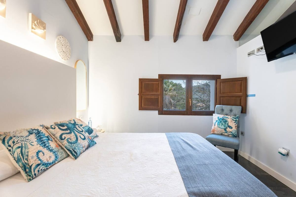 A bedroom with a bed, a chair, a window, and wooden beams on the ceiling.