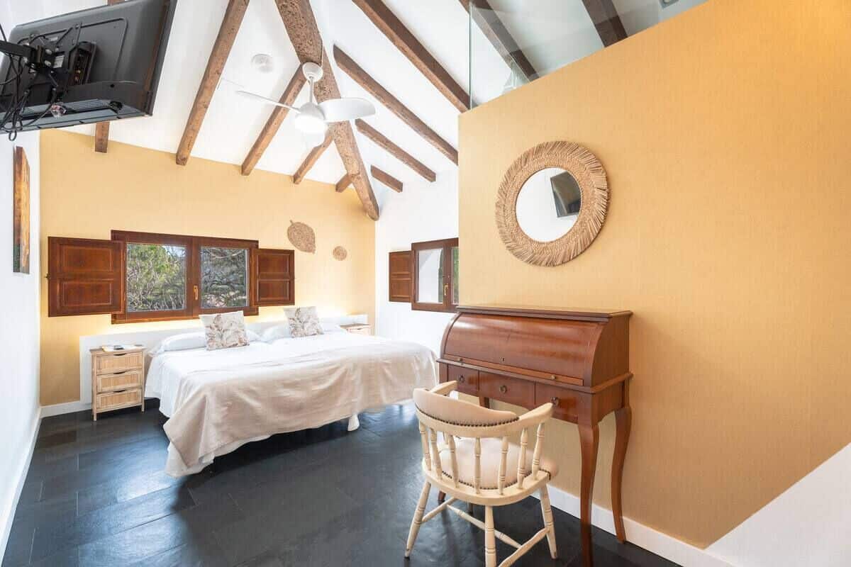 A bedroom with a bed, a wooden dresser, a chair, a mirror, and a television mounted on the ceiling.