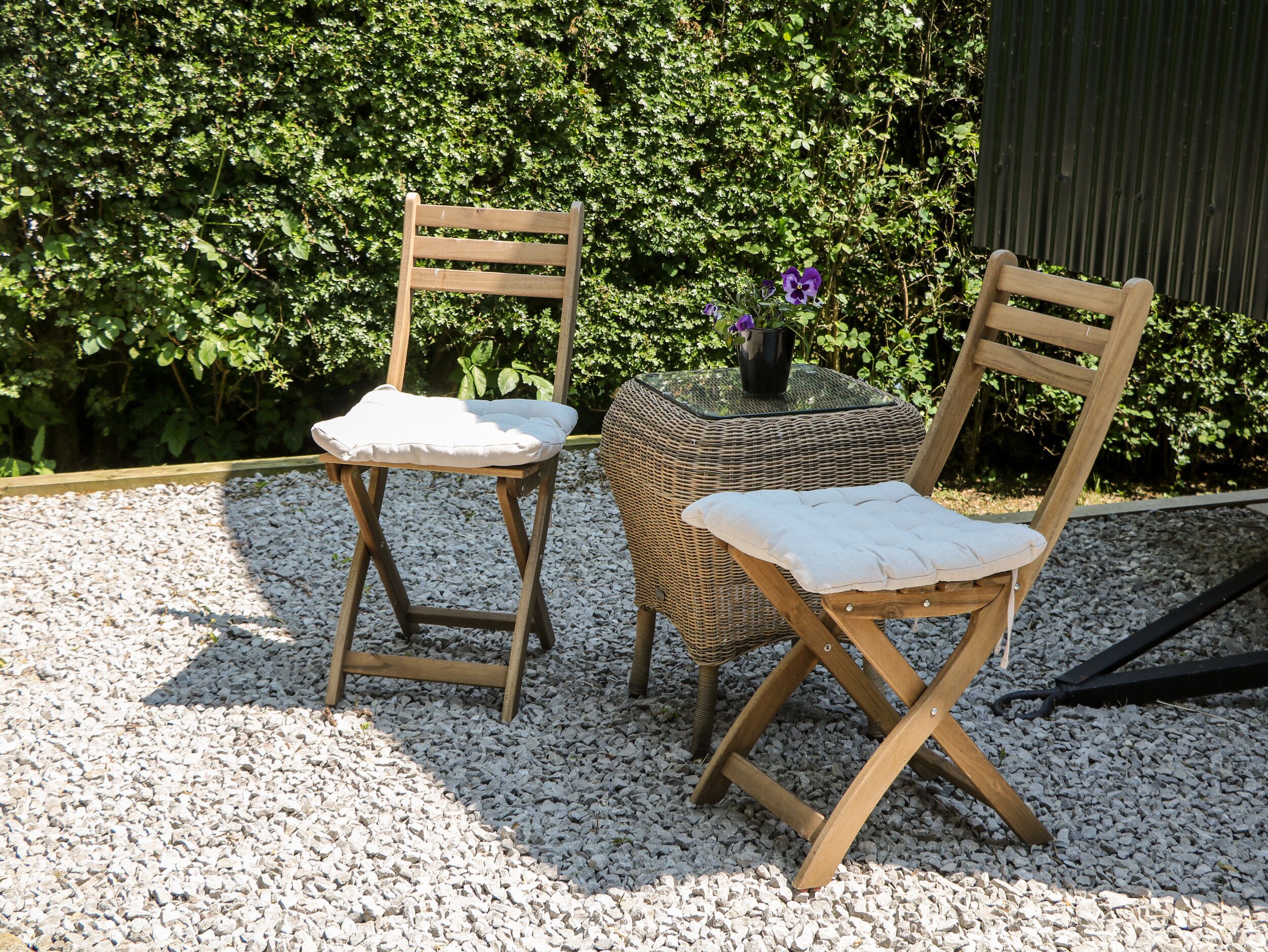 Twee houten stoelen met gewatteerde zittingen en rugleuningen, geplaatst op een grindpad met een rieten tafeltje ertussen.