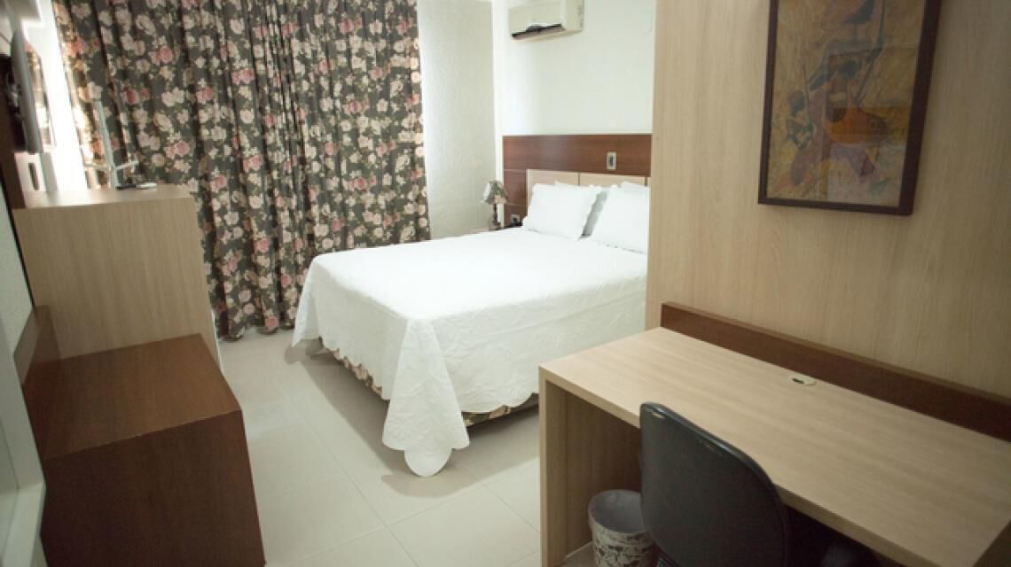 A hotel room with a bed, a desk, a chair, a trash bin, and a painting on the wall.