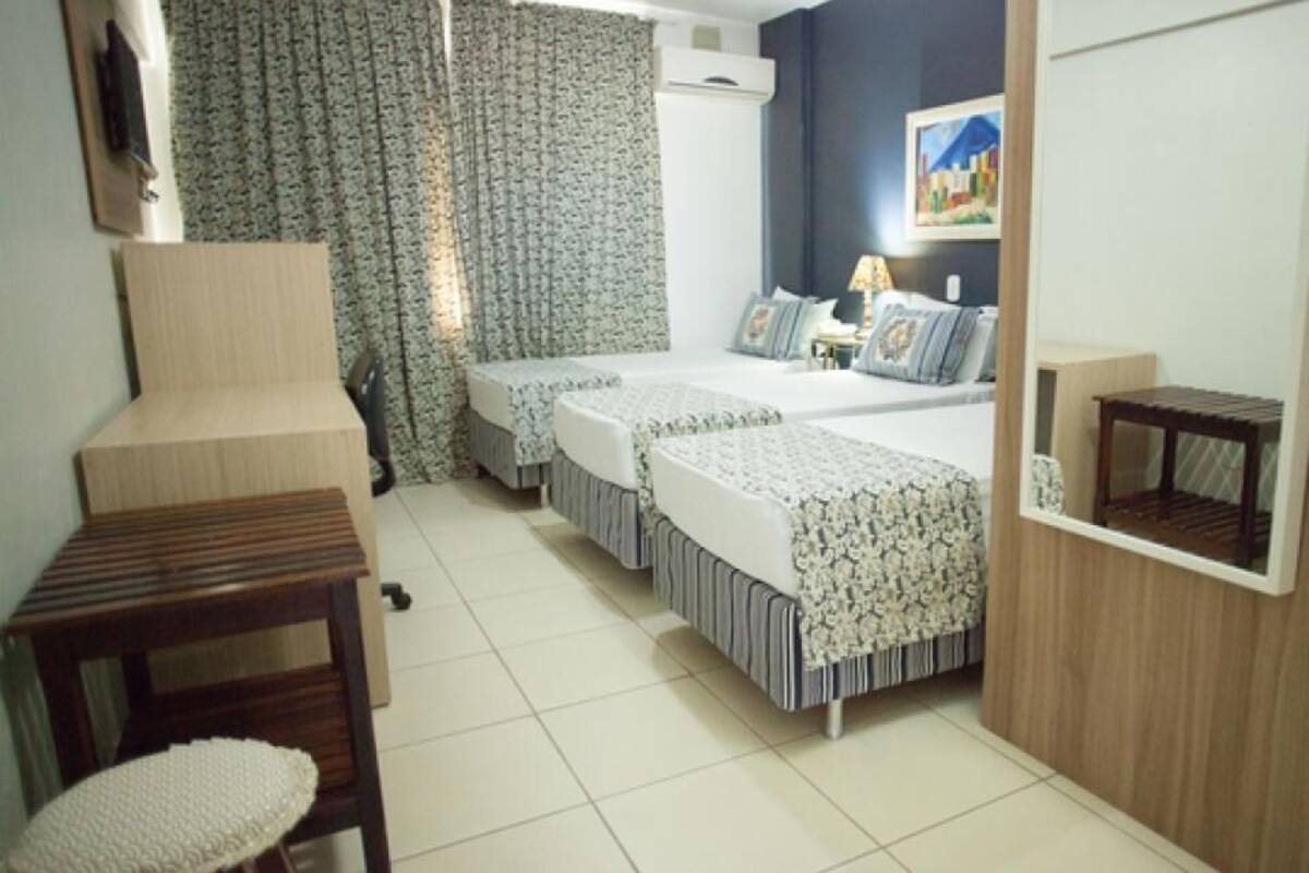 A hotel room with two beds, a wooden side table, a small stool, a wall-mounted TV, and a painting on the wall.