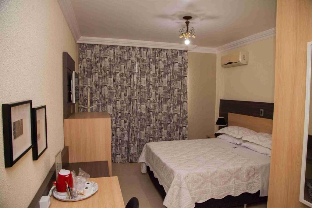 A small, neatly arranged bedroom with a bed, a desk, and a chair. There is a ceiling fan, a wall-mounted air conditioner, and a patterned curtain.
