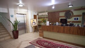 Un espace d’accueil avec un comptoir en bois, un portrait encadré, une plante en pot et un tapis rouge à motifs.