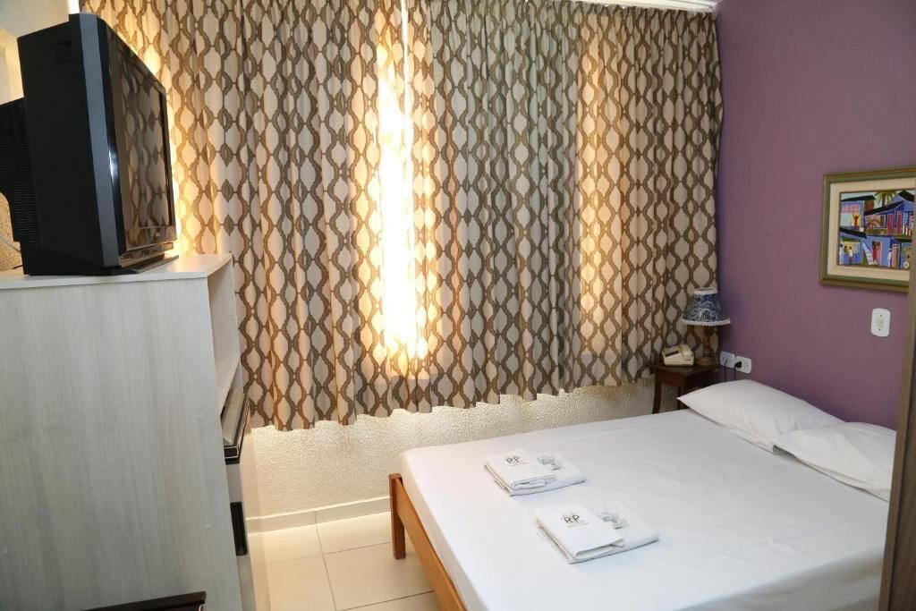 Une chambre d’hôtel avec un lit, un téléviseur posé sur un meuble, un rideau à motifs, une table de chevet avec une lampe et un tableau accroché au mur.