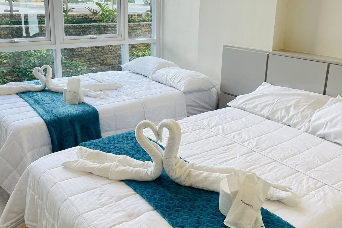 A hotel room with two beds, each with white linens and a teal blanket. There are two swan-shaped towels on each bed. The room has large windows with a view of the beach and palm trees.